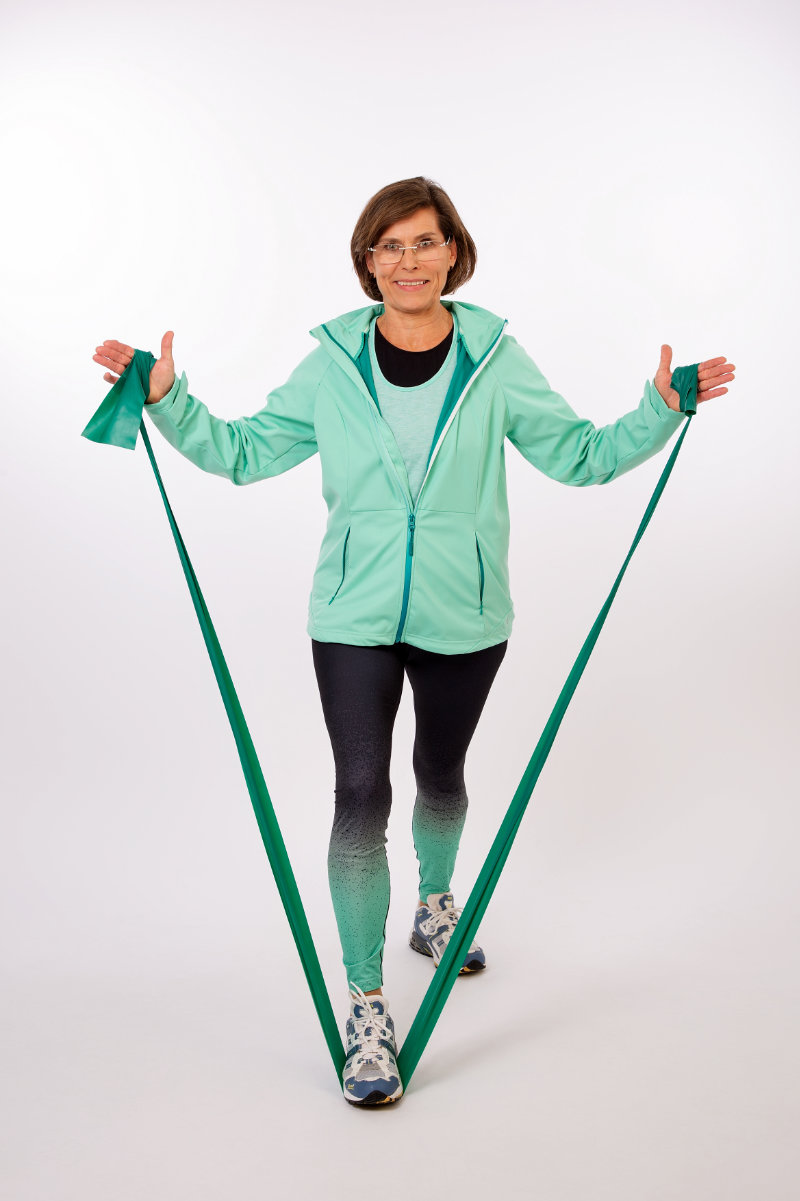 Margitta fixiert mit einem Fuß ein Band mittig am Boden und zieht die Enden mit jeweils einer Hand hoch